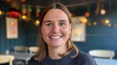 An image of Lucy Burns at a cafe in Eastbourne. Lucy has shoulder length brown hair and is wearing a navy jumper. She is smiling widely. 