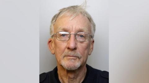 David Fretwell is wearing glasses with thin metal frames. He has wispy pepper and salt hair and has a white beard and a round face. He is wearing a black top.