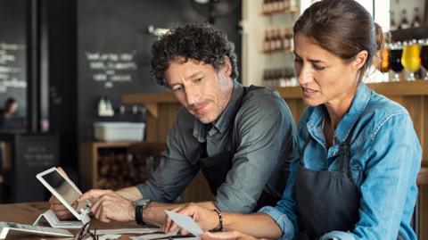 Two business owners - a man and a woman - sitting at a table in a bar, looking at receipts and holding a tablet