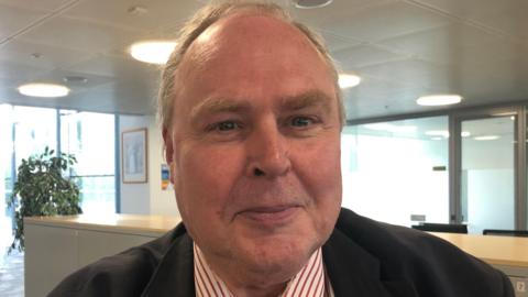 A photo of councillor Richard Smith. He is facing the camera and smiling. He is wearing a black suit with a red and white striped shirt underneath. Behind him is an office-style room. 