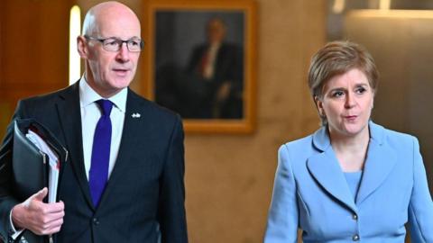 A bald man with glasses and a woman with short brown fair hair stand side by side. The man is wearing a dark suit, white shirt and blue tie. The woman is wearing a light blue suit. There is a painting on a wall behind them, out of focus in the back of the shot. 
