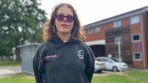 A woman stands looking at the camera. She has long brown hair and is wearing purple sunlgasses and a grey hooded sweatshirt. Behind her are brick buildings, parked cars and grass