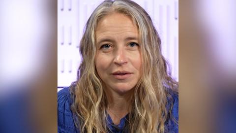 A woman with long blond wavy hair wearing a blue top, looks at the camera