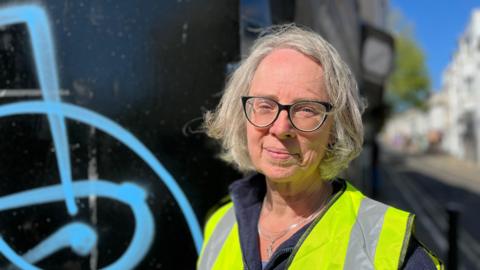 Emma Humphreys standing in Trafalgar Street, Brighton, next to a wall with blue spray paint on it. She has grey hair and is wearing a high vis vest