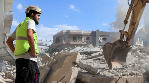 A rescuer in Lebanon, wearing a hard hat and high vis vest, has his hands on his hips and looks at an excavator at work on the rubble from a building hit by Israeli air strike