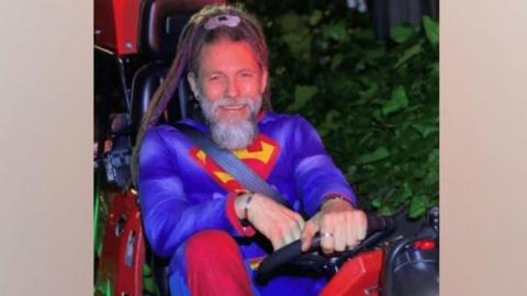 A man with grey hair pushed away from his forehead and a grey moustache and beard is wearing a Superman costume which is blue and red with a logo on the front. He is smiling into the camera and sitting in what looks like a go-kart, holding the wheel.