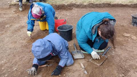 Three people are on their knees and crouched over as they investigate the soil below them. They are knelt in a shallow hole that has been dug in the field. They are all wearing blue waterproof jackets and gloves while archaeology equipment like spades and spatulas are strewn around them.