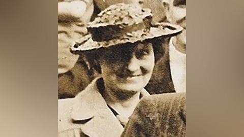 Louisa Dunne in the 1940s, stood among a crowd in a black-and-white photo. She is wearing a formal coat and brimmed hat, and is smiling.