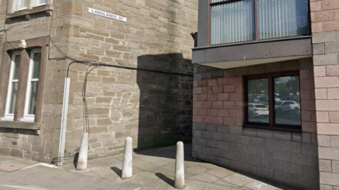 Langlands Street is a narrow lane with three concrete bollards at the entrance to it. There are buildings either side.