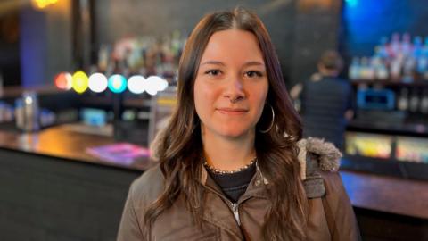 A woman wearing a brown jacket is standing in a bar. She has brown hair and a nose ring as well as large hoop earrings.