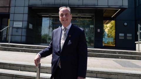 Councillor Ian Thorn stands outside office buildings with his hand resting on a railing. He smiles at the camera and has short grey hair. He is wearing a dark-blue suit jacket, a white shirt, and a white printed tie.