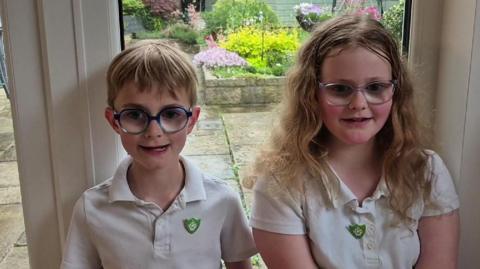Two children wearing glasses and white polo shirts with green Blue Peter badges on them.