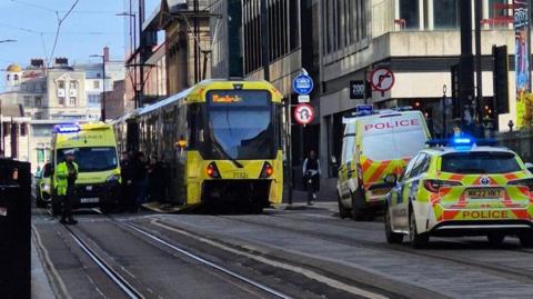 A tram stopped in the middle of the road, the front panel is missing. A police van and car are parked on the right side of the road. An ambulance is stopped next to the tram on the left. An officer wearing hi vis is stood in front of the ambulance.