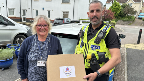 Mrs Wale and Sgt James Gallienne are holding a compassionate box. They are standing outside. Sgt James Gallienne is wearing a hi-vis police jacket. 