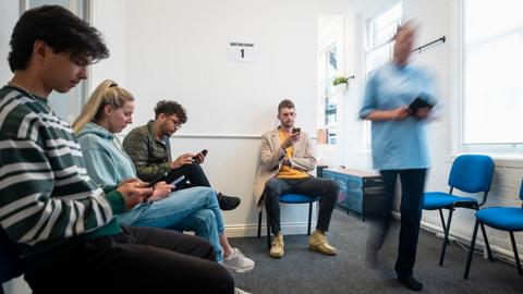 People wait in a healthcare waiting room as a healthcare professional walks though