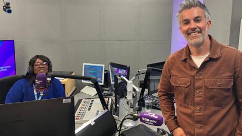 A woman, to the left of the image, sitting in a radio studio behind a microphone. She is wearing headphones and a blue jumper. A man is standing to her right. He is smiling and wearing a brown shirt.