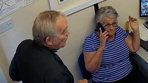 Two adults in a small office space, one seated and one standing, engaged in conversation. Papers and a computer monitor are visible in the background.
