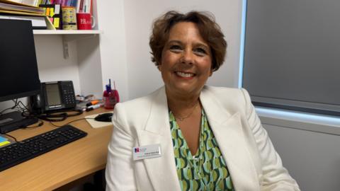 A woman with short brown hair smiling wearing a white blazer and a patterned light green, dark green and aqua blue top underneath. She is sat at a work desk with blinds closed behind.