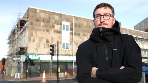 Jack Johnson at the High Street and Charles Road crossing. He has short brown hair, a moustache and glasses. He is wearing a black jacket and is standing with his arms folded. A traffic light can be seen behind him as well as traffic cones and a building with scaffolding erected at the front.