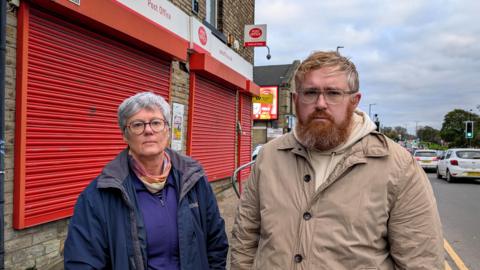 Two people standing in front of a post office branch with the shutters down. They both look very serious. The branch is on a busy road with lots of traffic passing. The man is middle aged and has ginger hair and a beard. He wears a beige jacket. The woman is older and had cropped greying hair. She wears a blue zip up jacket.
