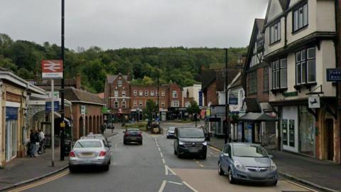 Station Avenue in Caterham has the railway station on one side and shops and residential accommodation nearby. There are cars on the road and people waiting outside the station, with countryside in the background.