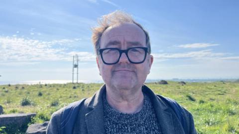Tony Lovell is wearing a grey and black knitted jumper underneath a grey jacket. He is standing in front of a field of grass with the sea on the horizon.