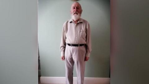 Patrick Williams in a light pink/white shirt and trousers. He is 5ft 7ins, of a slim build, with white/grey hair, a beard,