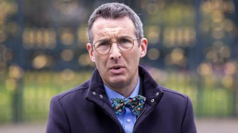 Andrew Muir looks into the camera. He is wearing round glasses, a navy overcoat, blue shirt and a brightly patterned bow tie. He has black and grey hair. The black and gold gates of Hillsborough Castle are in the background.