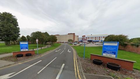 A road leading to the entrance of Hartlepool Hospital. There are two blue signs and on one of them it says there is no A&E. The hospital s in the background and is spread across two or three buildings.