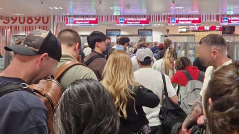 A crowd of passengers waiting in the passport control queue at Milan Bergamo airport on 16 April, with four check-in desks visible.