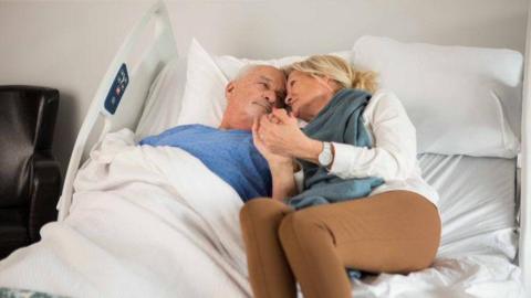 An elderly couple in a wide hospital bed, cuddling and smiling at each other.