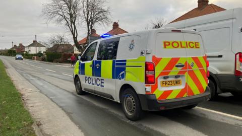 Police vehicle at the scene of the incident in Hornsea. There are trees and houses in the background with a blue car on the opposite side of the road.