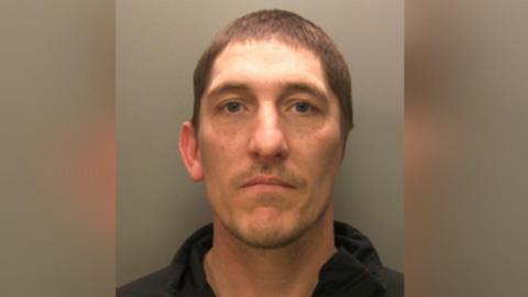 A mugshot of Christopher Hewer. He has short brown hair and blue eyes and his wearing a black hoodie. He only has one ear.