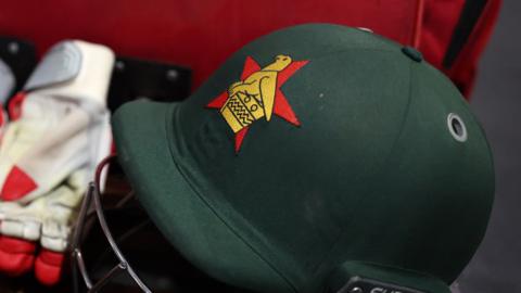 A green cricket helmet with a red and gold Zimbabwe cricket crest is visible, with a white and red batting glove sitting on the left-hand side of the image up against the helmet's grill