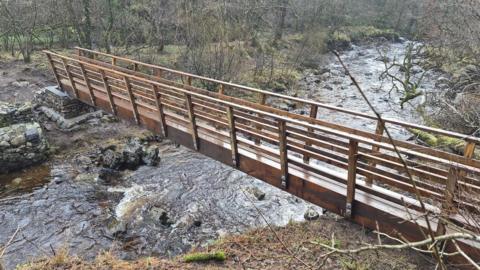 A wooden footbridge with stone steps spanning a small river in full flow. The scene is wintry with a background of woodland with leafless trees.