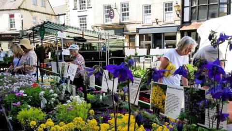 A group of people browsing a flowers and plants stall in a street market. In the foreground are yellow and blue flowers, with buildings and other market stalls in the background.