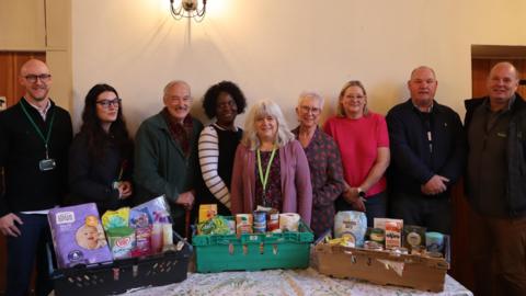 A group of people stand in a row with a table in front of them. On the table are boxes with essential items in them, such as nappies, food and milk.