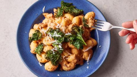 Veggie sausage gochujang gnocchi with crispy kale in a blue bowl on a pale grey background. 