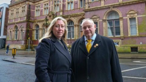 A woman with long blonde hair and a man with short greying hair are both wearing long black coats. They are standing in a city street in front of an ornate brick building with honey-coloured arched windows throughout.