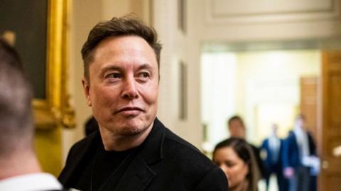 Elon Musk wearing a dark jacket. He is looking away from the camera with a smirk on his face