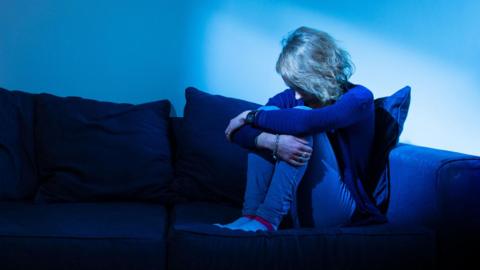 A woman huddled up with her face down and not visible on the end of a sofa. The image is sepia tone with a shadow over the couch with just a small amount of light over her head. 