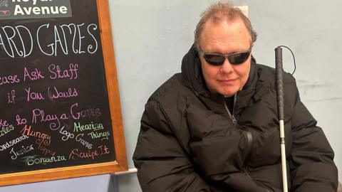 Dave Cunningham is wearing dark glasses and a black coat, and has a white cane in his hand. He is sitting sitting against a grey wall. There is a blackboard on one side and a bookcase on the other.
