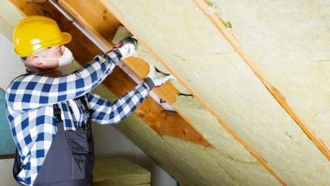 Man in a yellow hard hat, checkered shirt and overalls insulating a house 