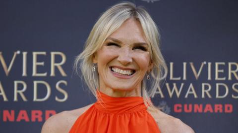 Jo Whiley poses on the red carpet at the Olivier Awards, wearing a bright orange sleeveless dress in front of an event backdrop featuring the Olivier Awards and Cunard logos.
