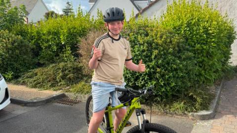A boy on a push bike is smiling at the camera. He is wearing a safety helmet and is doing the thumbs up sign with both hands. He is in front of a large green hedge an appears to be in a car park near some houses. 