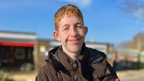 17-year-old Aidan smiling at the camera. He has short auburn hair and is standing outside on a sunny day, and there is a school building in the background. 