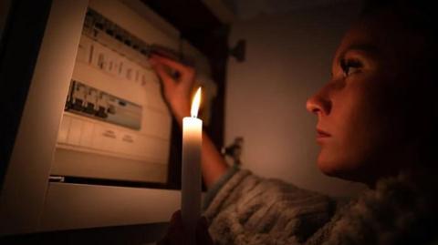 A woman in a dark room looks at a fusebox by candlelight