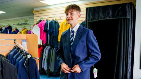 A teenage boy with short brown hair, standing in a clothes shop. He is wearing a navy blazer, white shirt and stripy blue tie. 