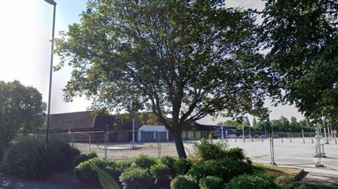 A boarded up supermarket building with barriers around the car park. The site stands empty. There are trees and shrubs planted around the site.