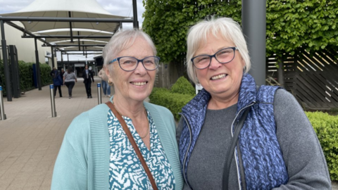 Two women with glasses, smile. The woman on the left, with white hair wears a turqoise cardigan. The woman on the right with white hair wears a grey jumper and blue gilet.